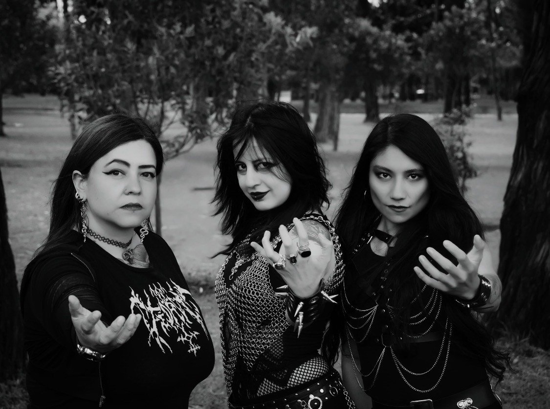 Three women dressed in gothic style posing outdoors in a forested area.