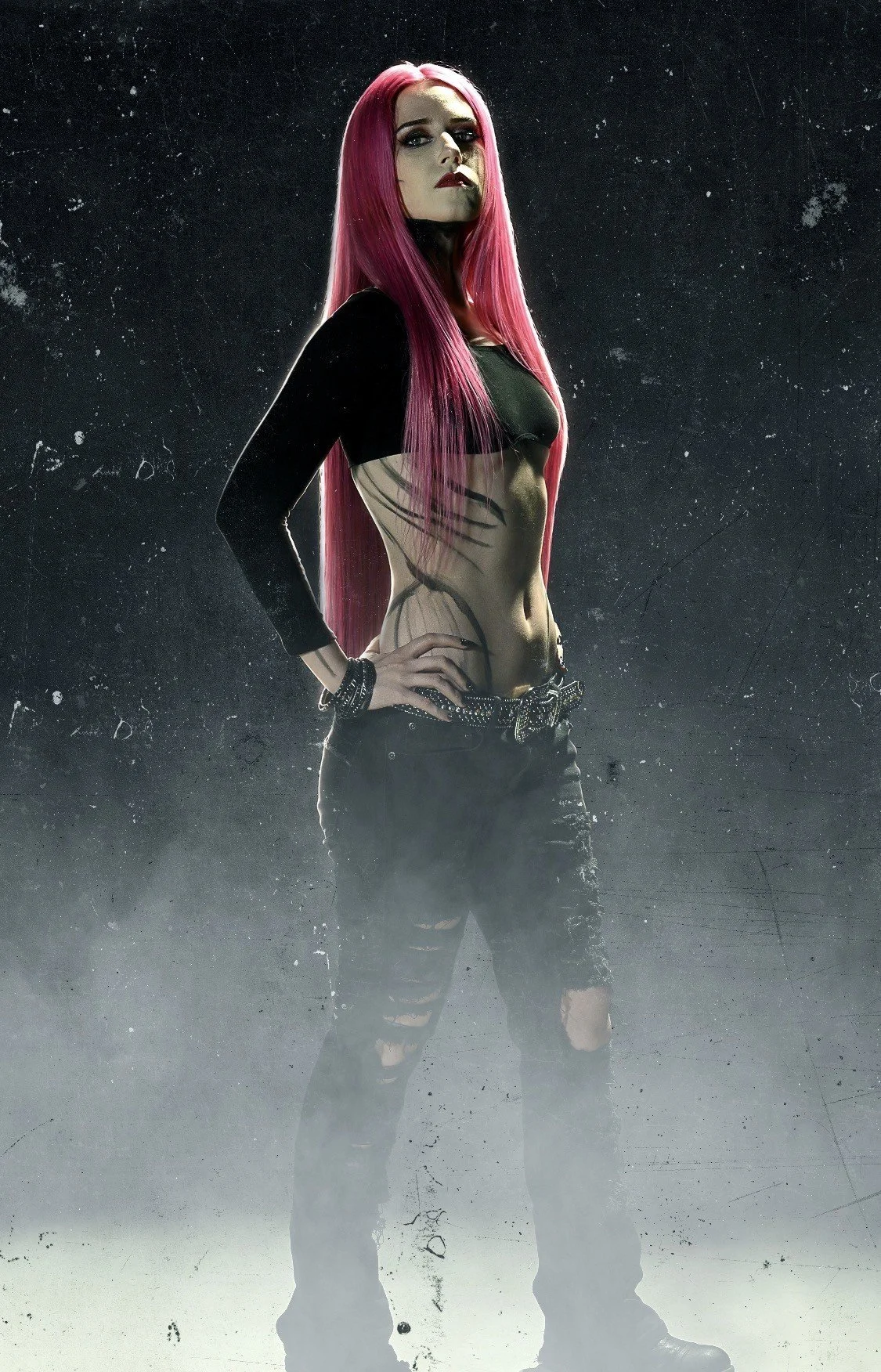 A woman with long pink hair, wearing black ripped jeans and a black top. She has dark makeup and body paint resembling a spider on her torso. She stands against a dark foggy background with her hand on her hip.