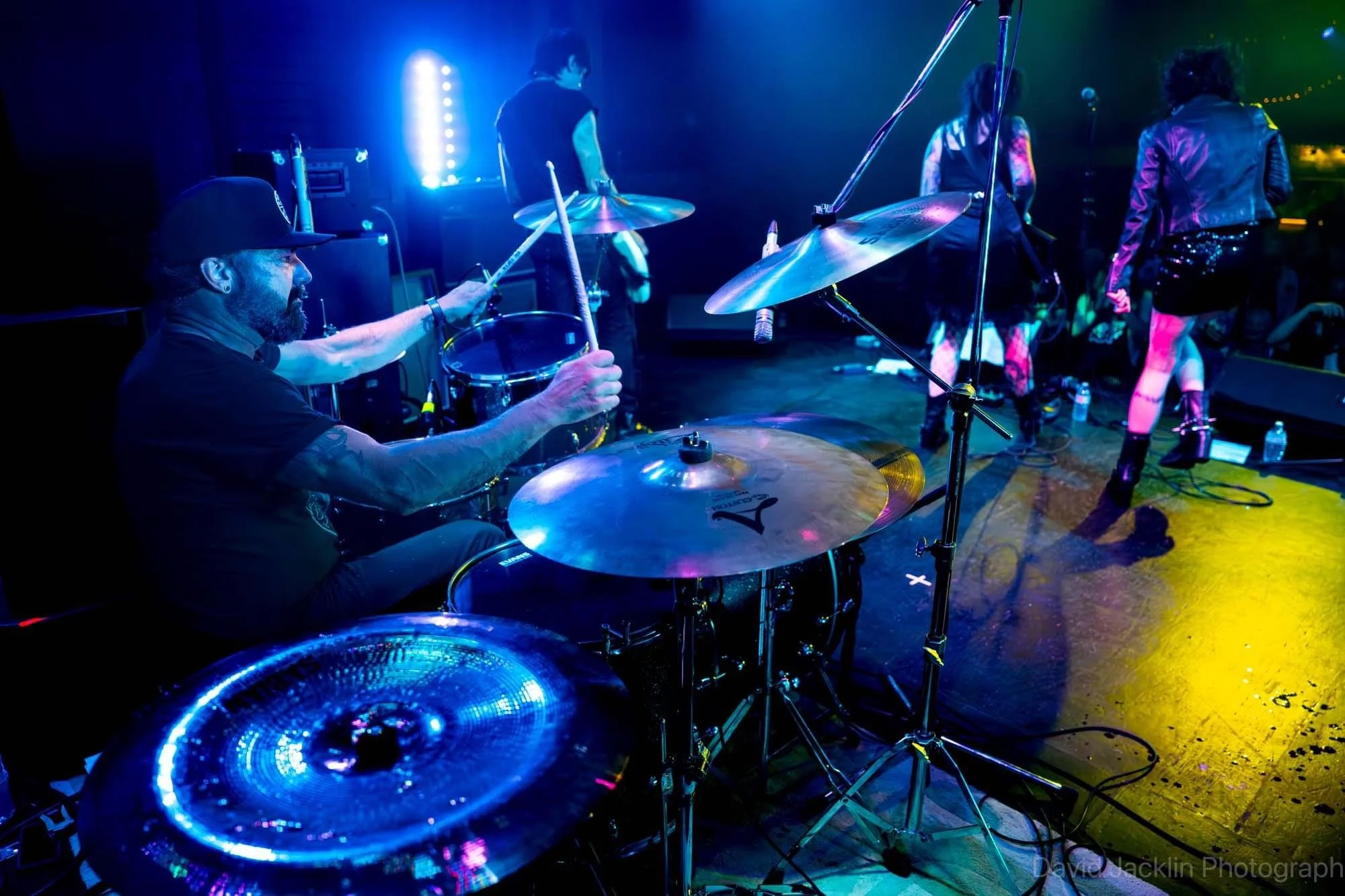 View of a band performing on stage with a drummer playing in the foreground, surrounded by colorful stage lights, and other band members visible in the background.