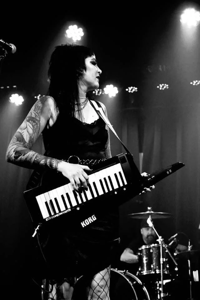 A female musician with short dark hair and tattoos playing a keytar on stage during a concert, with a drummer in the background.