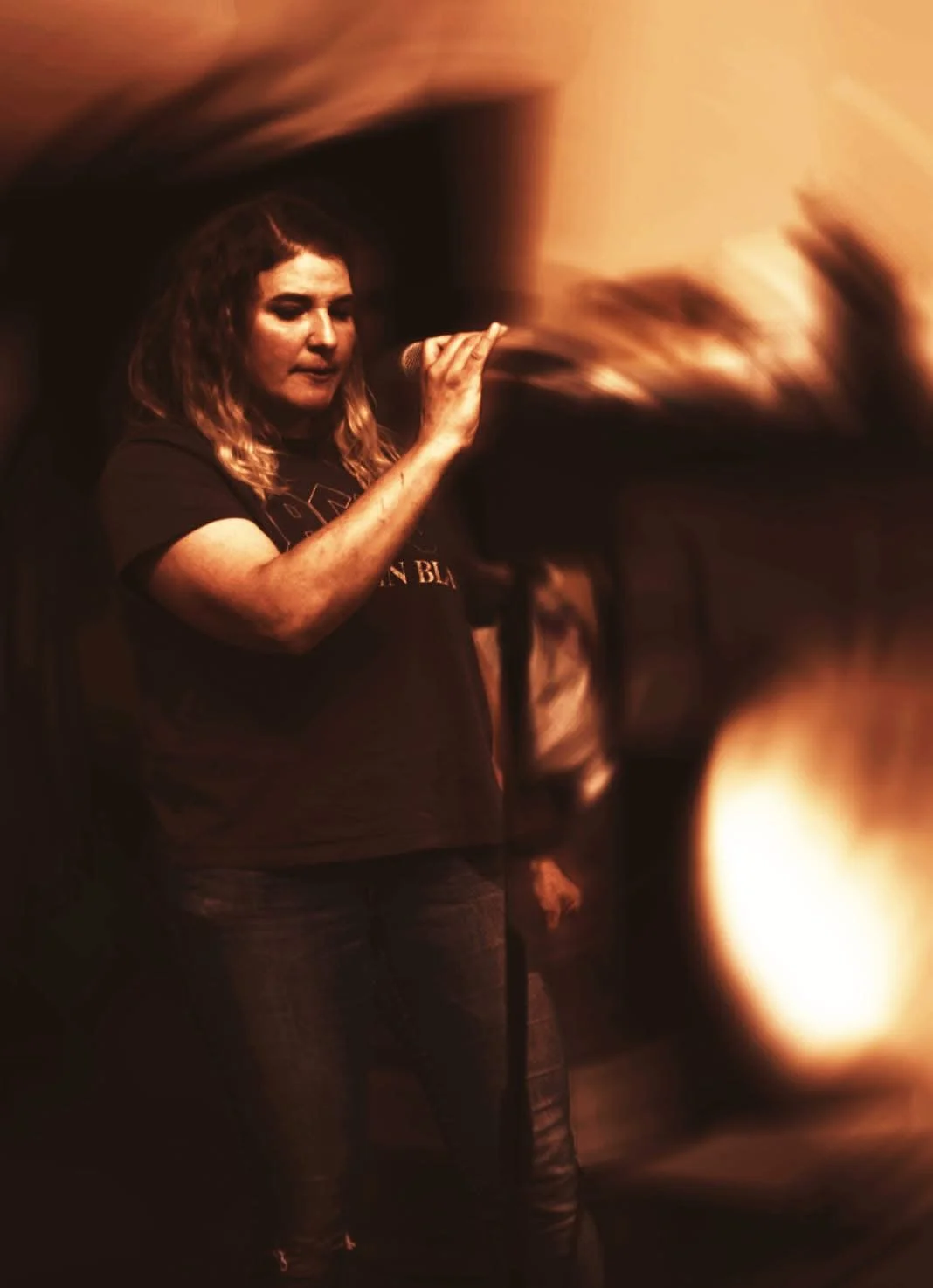 A woman with long hair holding a microphone in a dark, blurry environment, possibly performing or recording.