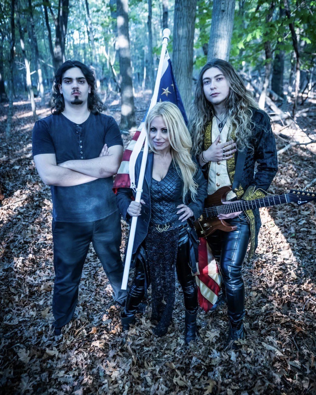 Three young adults standing in a wooded area, with one holding an American flag and another holding an electric guitar.
