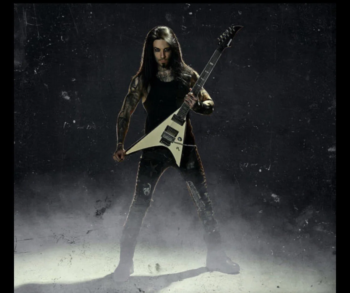 A woman with long dark hair, tattoos, and dark clothing standing in a smoky environment, holding a white electric guitar in a pose.