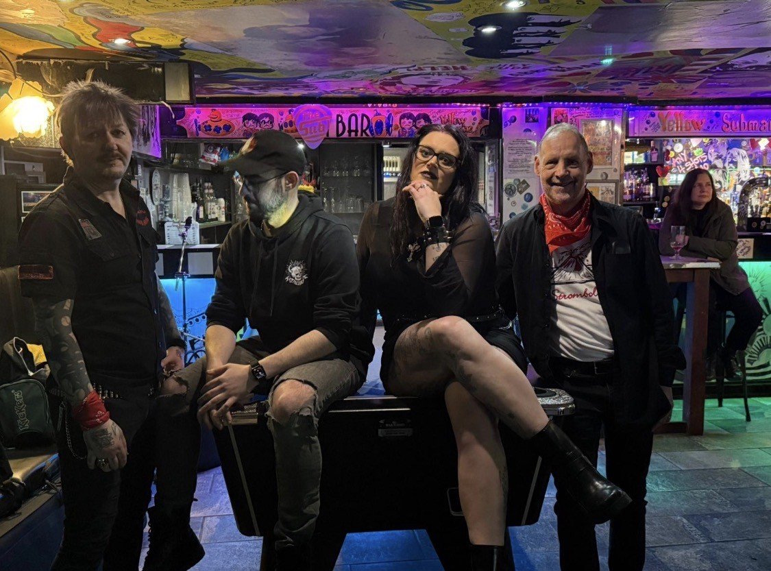 Five people in a bar or music venue, with colorful wall art and neon lighting, are posing for a photo. One person with face paint and a hat is sitting on a speaker, others are standing around, and one woman is sitting on the speaker with her leg crossed.