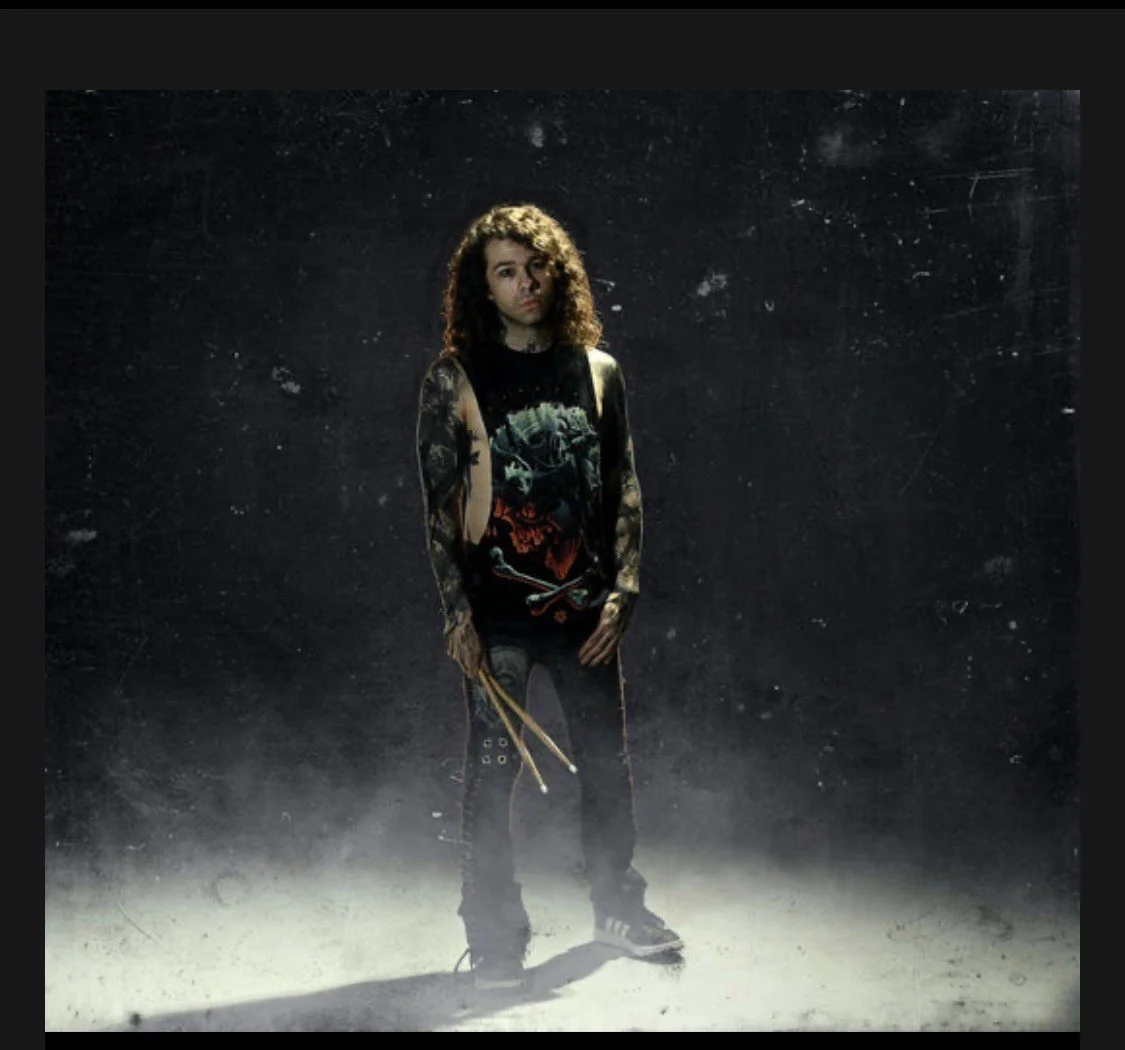 Young man with curly hair standing in a dark, smoky setting, wearing a sleeveless Iron Maiden shirt, ripped jeans, and holding drumsticks.