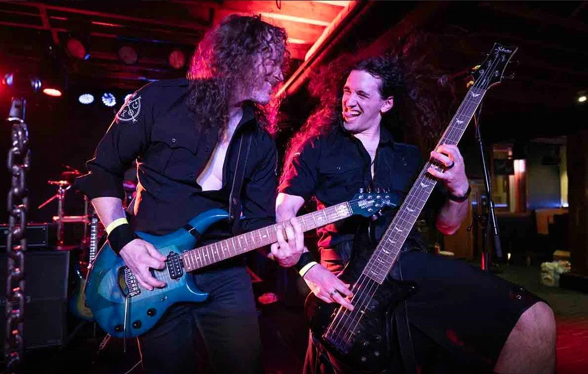 Two male musicians playing electric guitars on stage, smiling and enjoying the performance, with colorful stage lights in the background.