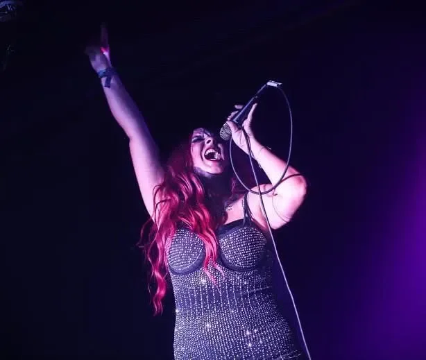 Female singer performing on stage with long red hair, wearing a sparkly black dress, holding a microphone, with one arm raised and expressive singing face.