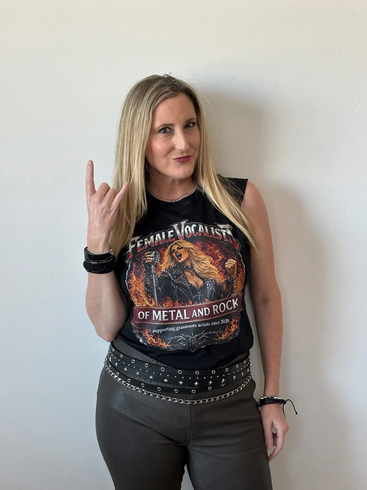 Woman with blonde hair in black sleeveless shirt, making a rock hand gesture against a plain white wall.