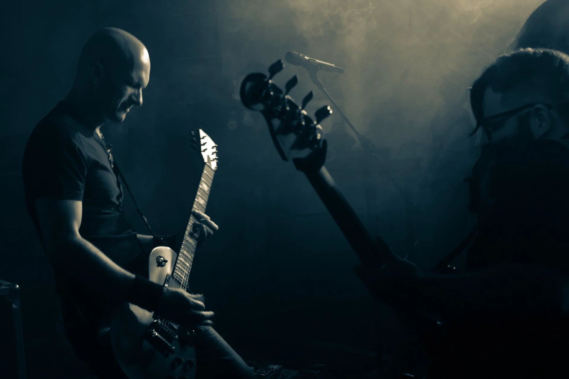 A musician playing an electric guitar in a dimly lit setting, with a microphone stand nearby, and another person with glasses slightly visible on the right.