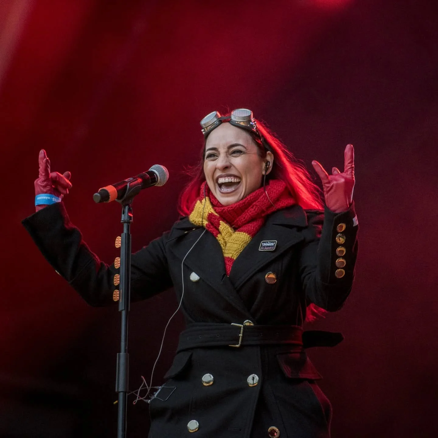 A woman with red hair, wearing goggles on her head, a black coat, a red and yellow scarf, and red gloves, standing in front of a microphone on a stage with a dark background, smiling and making hand signs.
