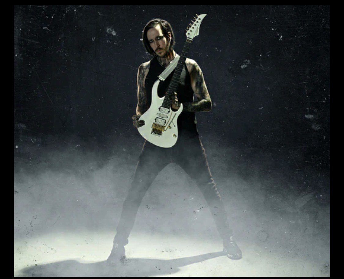 A person with black hair and tattoos on their arms, wearing a black sleeveless shirt and black pants, is standing in a smoky, dark environment, holding a white electric guitar with a black neck and a unique headstock.