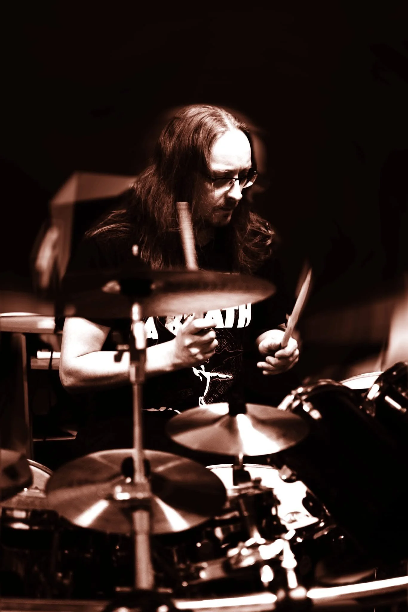 A man with long hair and glasses playing drums in a dark room, wearing a black t-shirt.