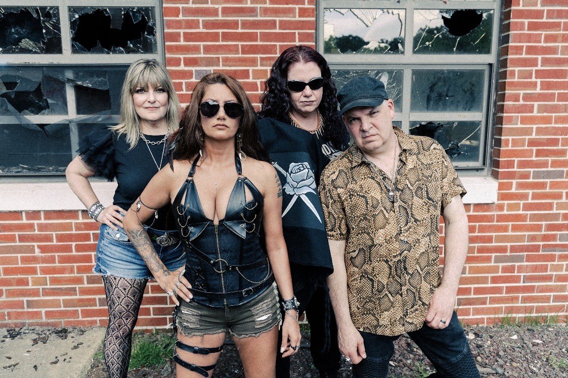 Four people standing in front of a broken window with a brick wall. They are dressed in edgy, punk-inspired clothing.