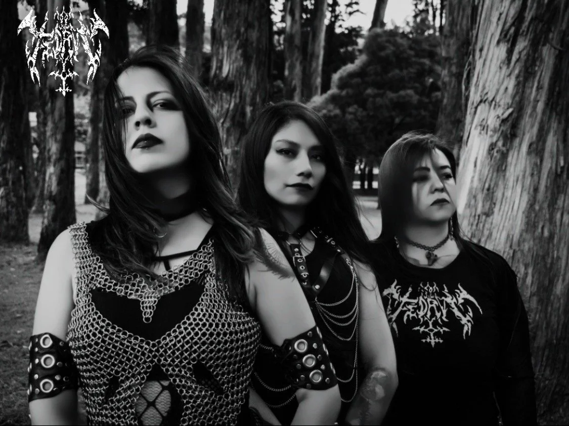 Three women with gothic style standing outdoors among trees in black and white photo.