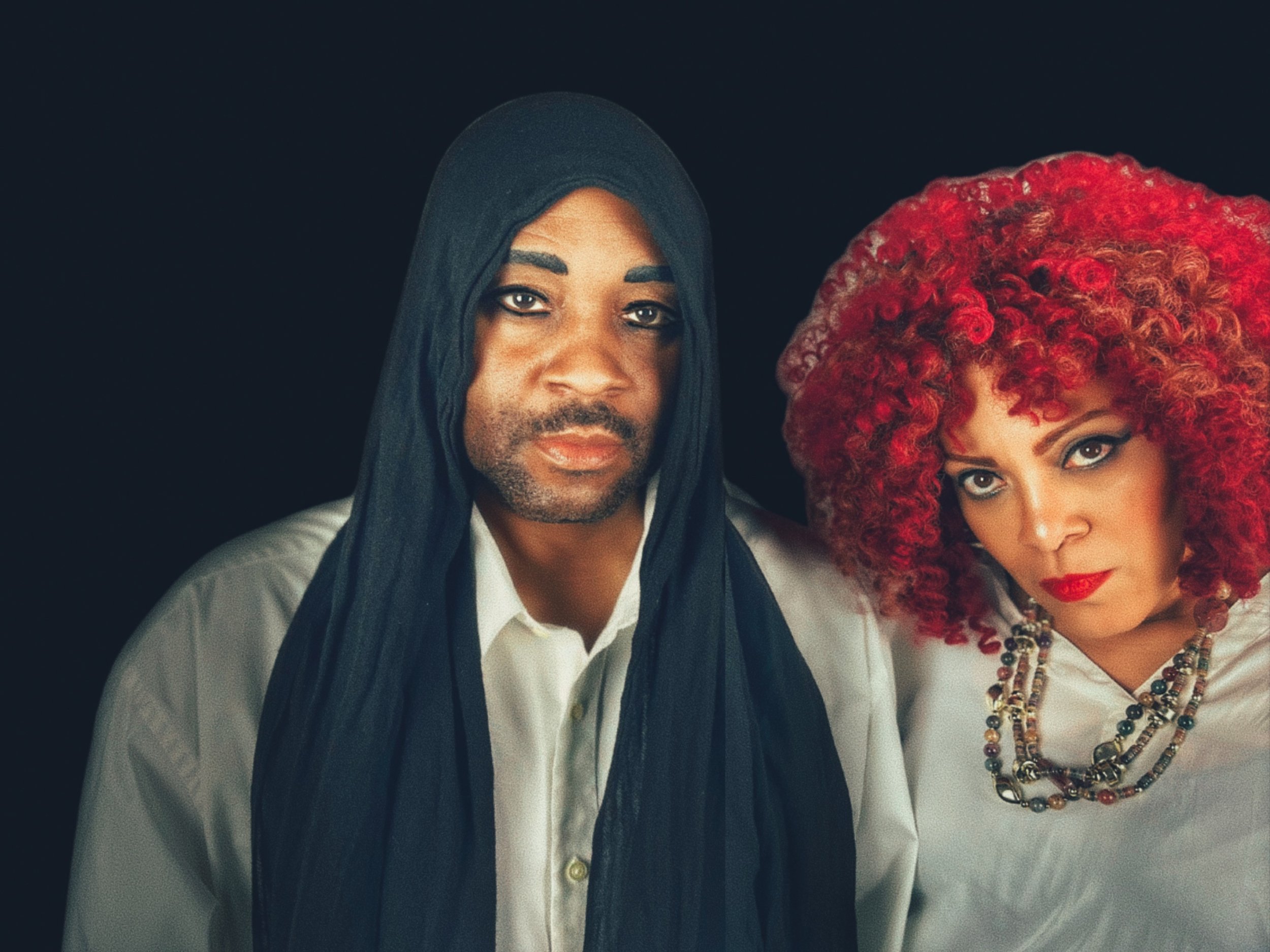 Close-up of a man wearing a black hijab and white shirt standing next to a woman with curly bright red hair, wearing a white shirt and multiple colorful beaded necklaces, against a black background.