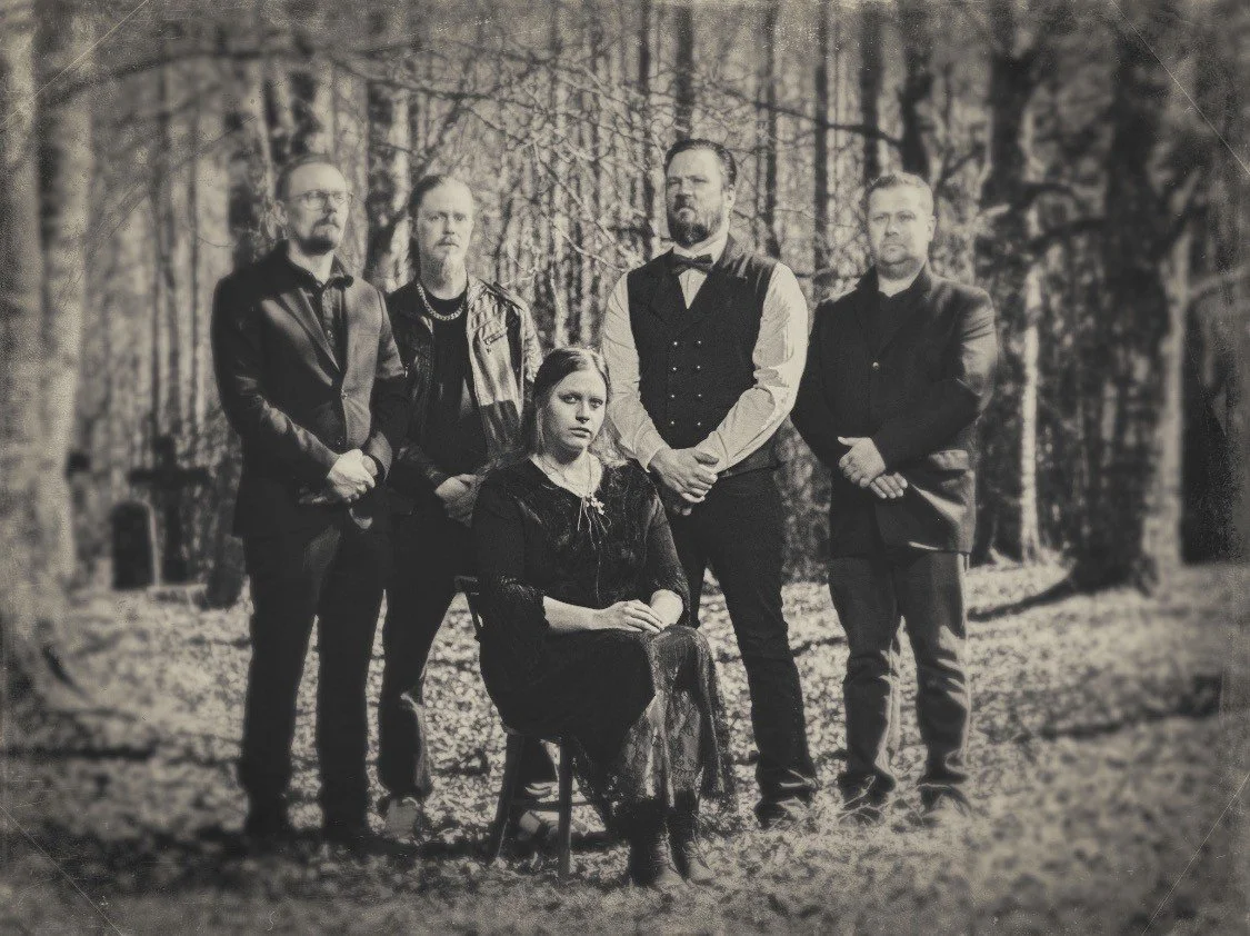 A black and white photograph of six people posing outdoors in a wooded area. One woman is seated in front, and five men stand behind her, all dressed in dark clothing.