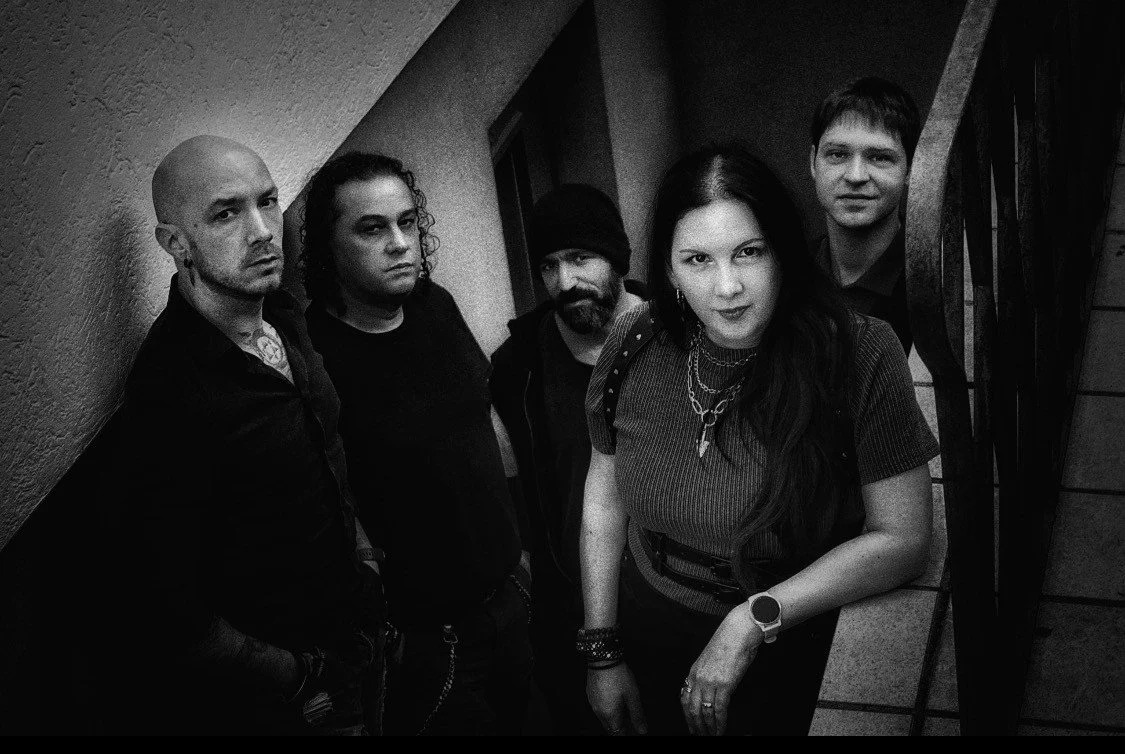 Five people standing on a staircase in a narrow hallway, facing the camera with serious expressions. The woman in the front has long dark hair and wears a striped top with layered necklaces. The man on the far left is bald with a tattoo on his neck, and the man on the far right has short hair. The woman second from the left has curly hair, and the man in the middle wears a beanie.