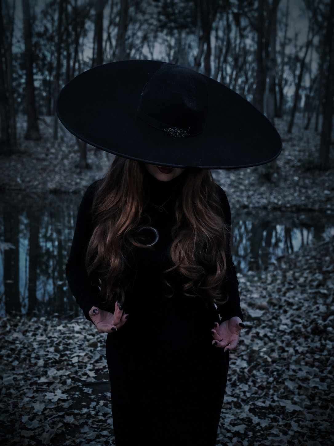 A woman in a dark dress and large black hat standing in a wooded area near a small body of water, with trees and fallen leaves visible around her.