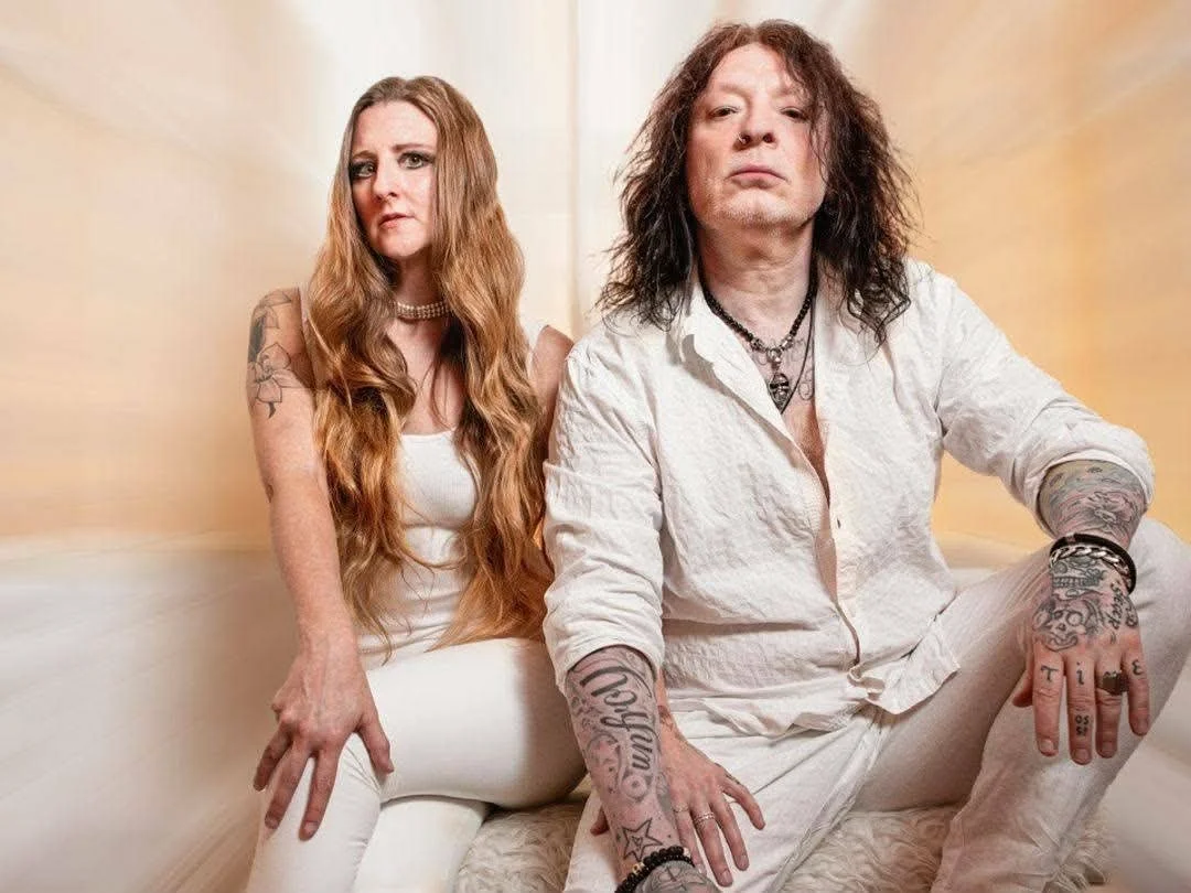 A woman with long red hair and tattoos on her arms sitting on a white bench next to a man with long dark hair and tattoos on his arms, both dressed in white, posing against a cream-colored wall.