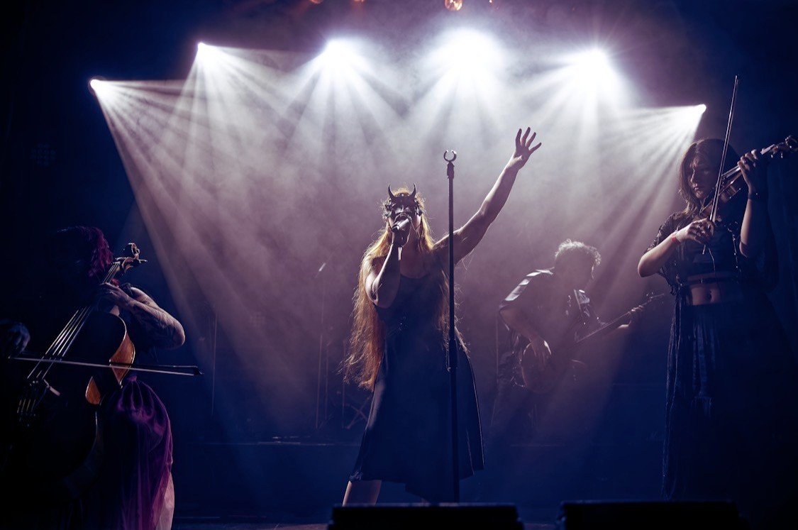A performer wearing a mask with horns singing into a microphone on stage with three other musicians playing instruments, under bright stage lights.