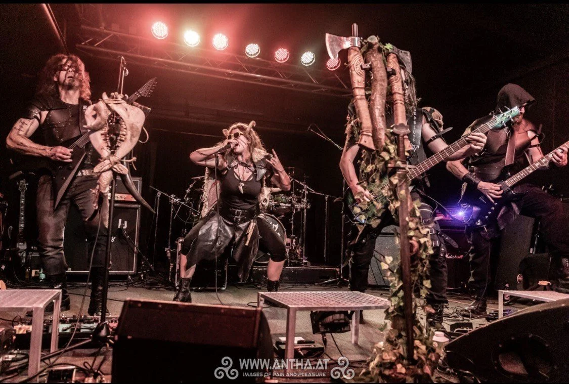 A band performs on stage with band members dressed in dark, gothic costumes, some wearing horns and masks, under red stage lights, with musical equipment and instruments visible.