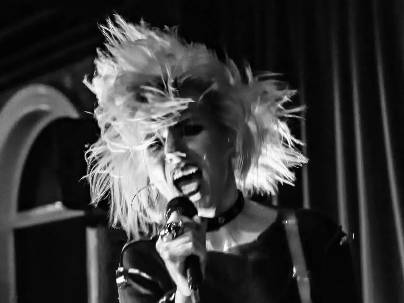 A person with wild, styled hair singing passionately into a microphone with dark background.