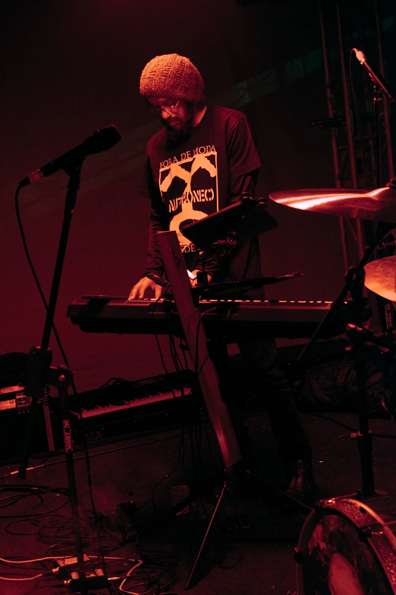 A man wearing a gray knit beanie, glasses, and a black t-shirt with white text and graphics is playing electronic music on a keyboard and electronic equipment in a dimly lit setting with red lighting. There are microphones, stands, and cables around him.