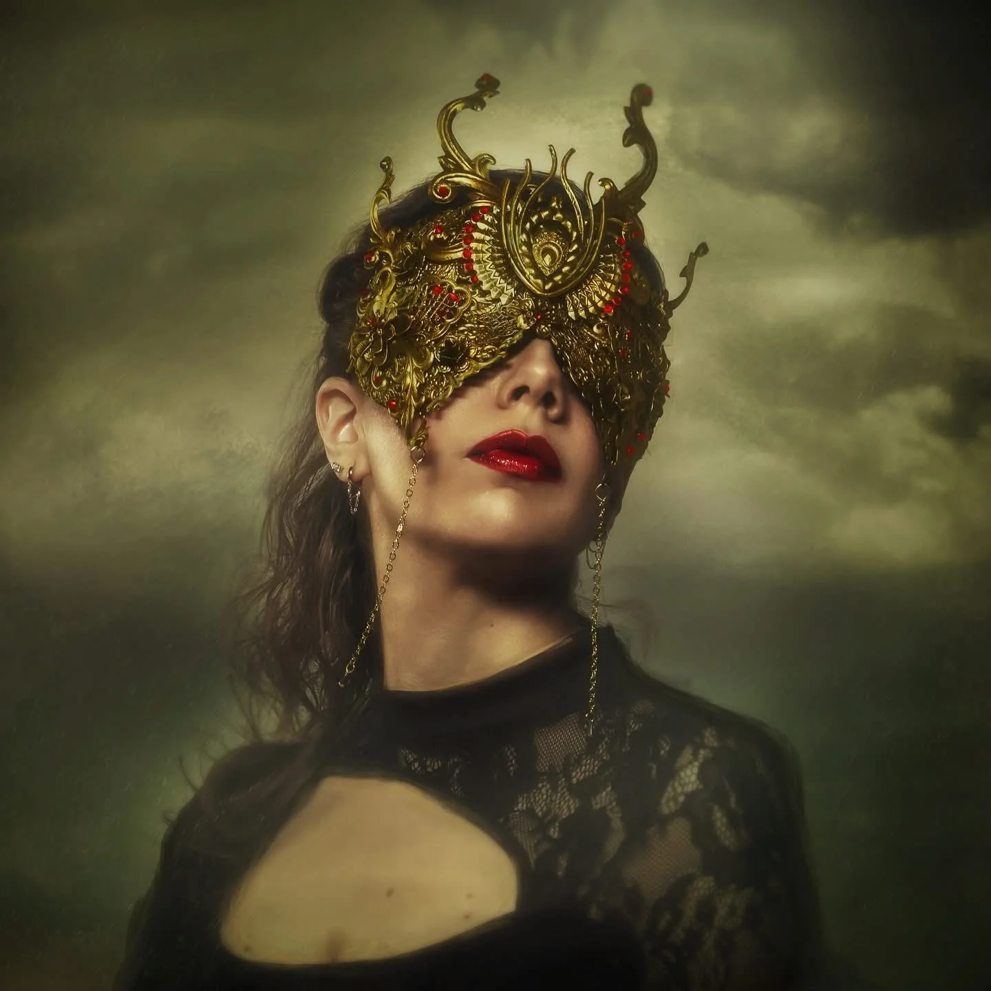 A woman wearing an ornate gold dragon mask covering her eyes, with dark curly hair, red lipstick, and earrings, dressed in black lace clothing, against a cloudy sky background.