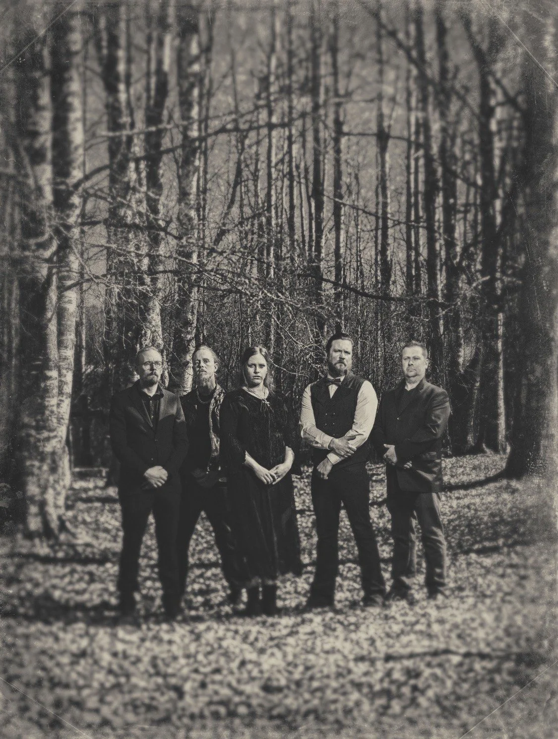 A black-and-white photo of five people standing in a forest during autumn, dressed in formal or vintage clothing.
