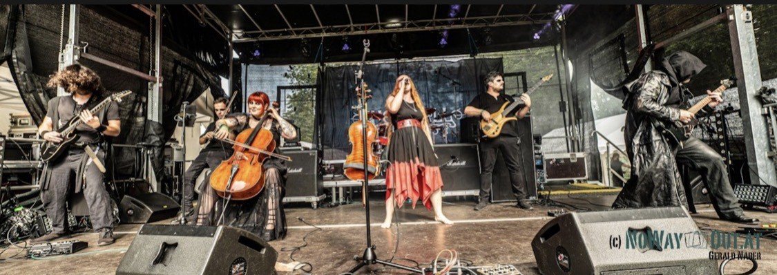Band performing on stage outdoors with seven musicians playing guitars, violin, and singing.