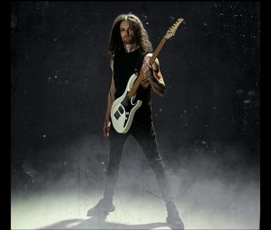 A man with long curly hair, wearing a sleeveless shirt, stands holding a white electric guitar against a dark, textured background with a foggy floor.