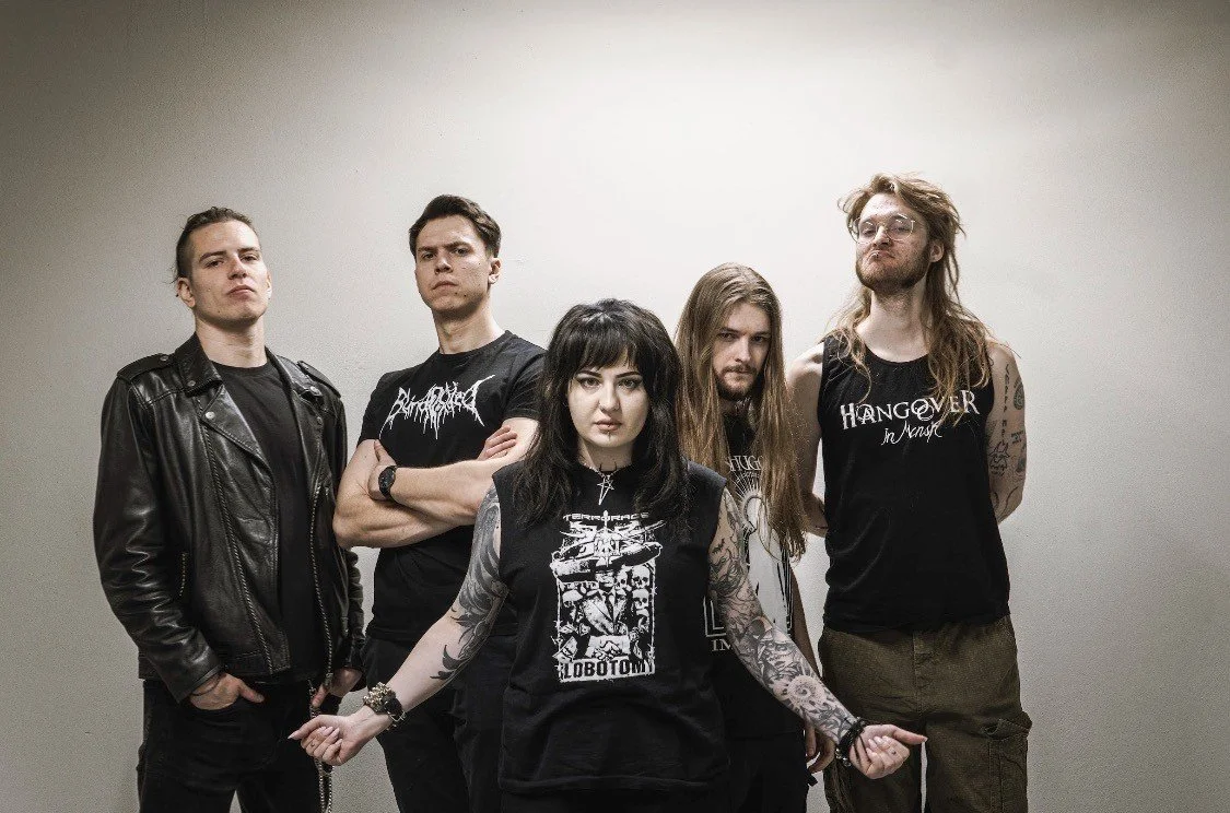 Group of five young adults posing against a plain wall, dressed in black clothing, with some wearing band T-shirts and displaying tattoos.