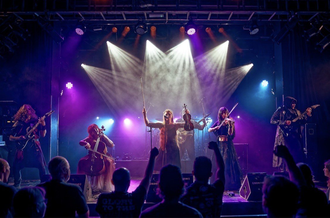 A band performing on stage with musical instruments like guitars, violin, and cello, under colorful stage lights, with an audience watching and raising their fists.