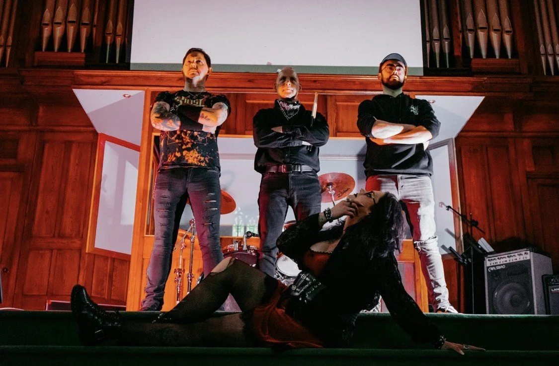 A woman with black hair, wearing a black jacket and red skirt, is lying on the stage floor in front of a band, posing with her head tilted back and her hand on her chin. The band members behind her are three men with crossed arms and serious expressions, standing on a wooden stage in a music venue or recording studio.