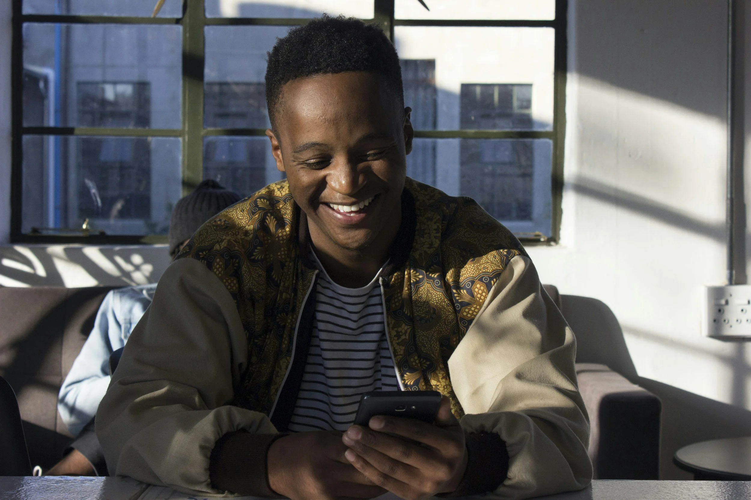 A young man with short black hair smiling and looking at his phone, sitting at a table indoors with sunlight streaming through large windows behind him.