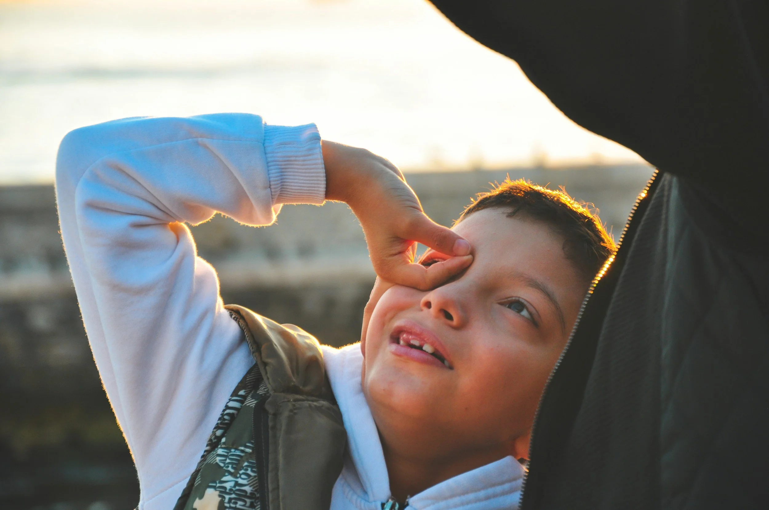 A young boy wearing a white hoodie and a black vest with a pattern is smiling and making a playful gesture with his hand near his eye, while an adult's arm is visible holding his other hand. The background shows a blurred outdoor setting with sunlight.
