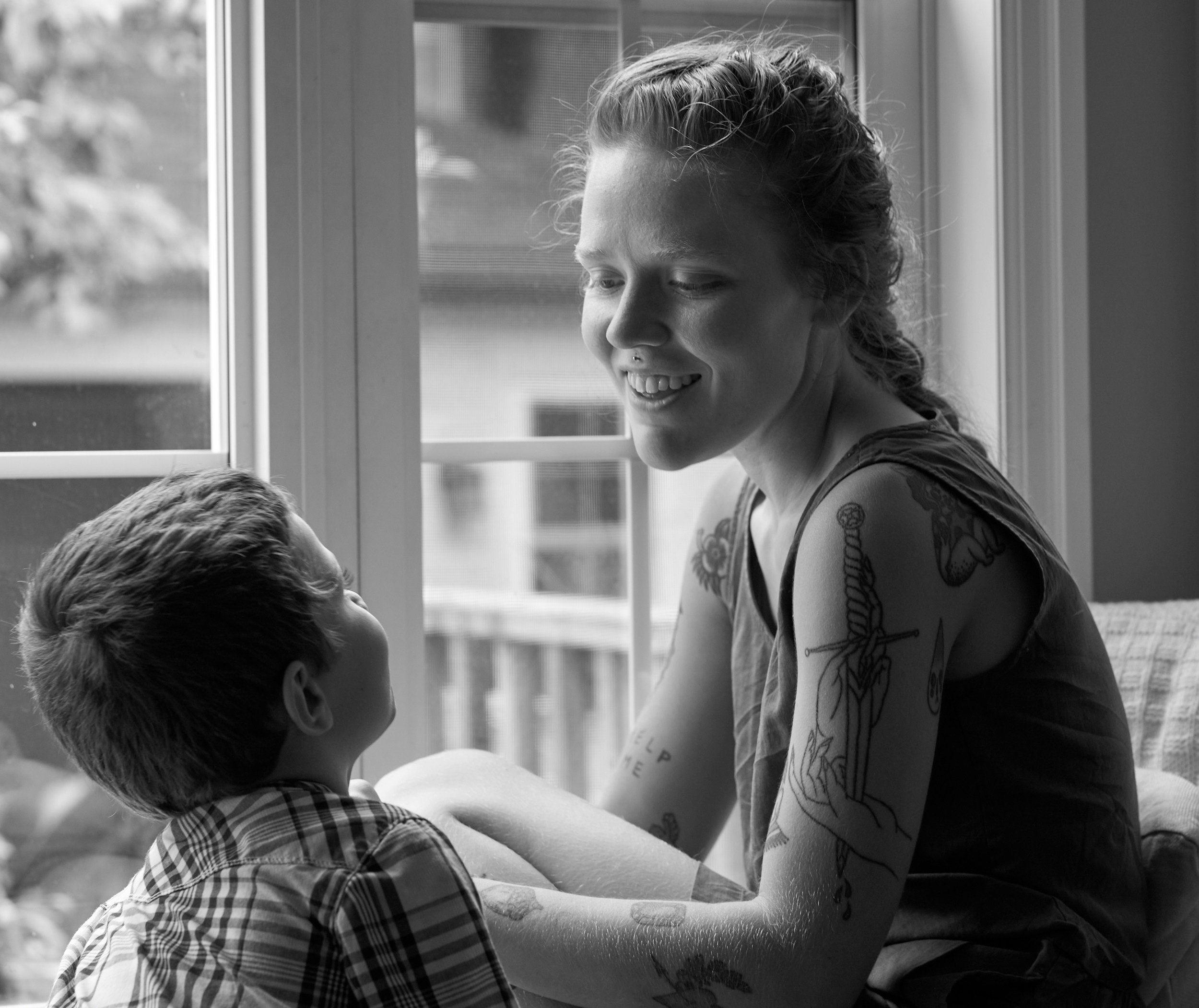 A woman with tattoos on her arm smiling and talking to a boy with short hair and a checkered shirt by a window.