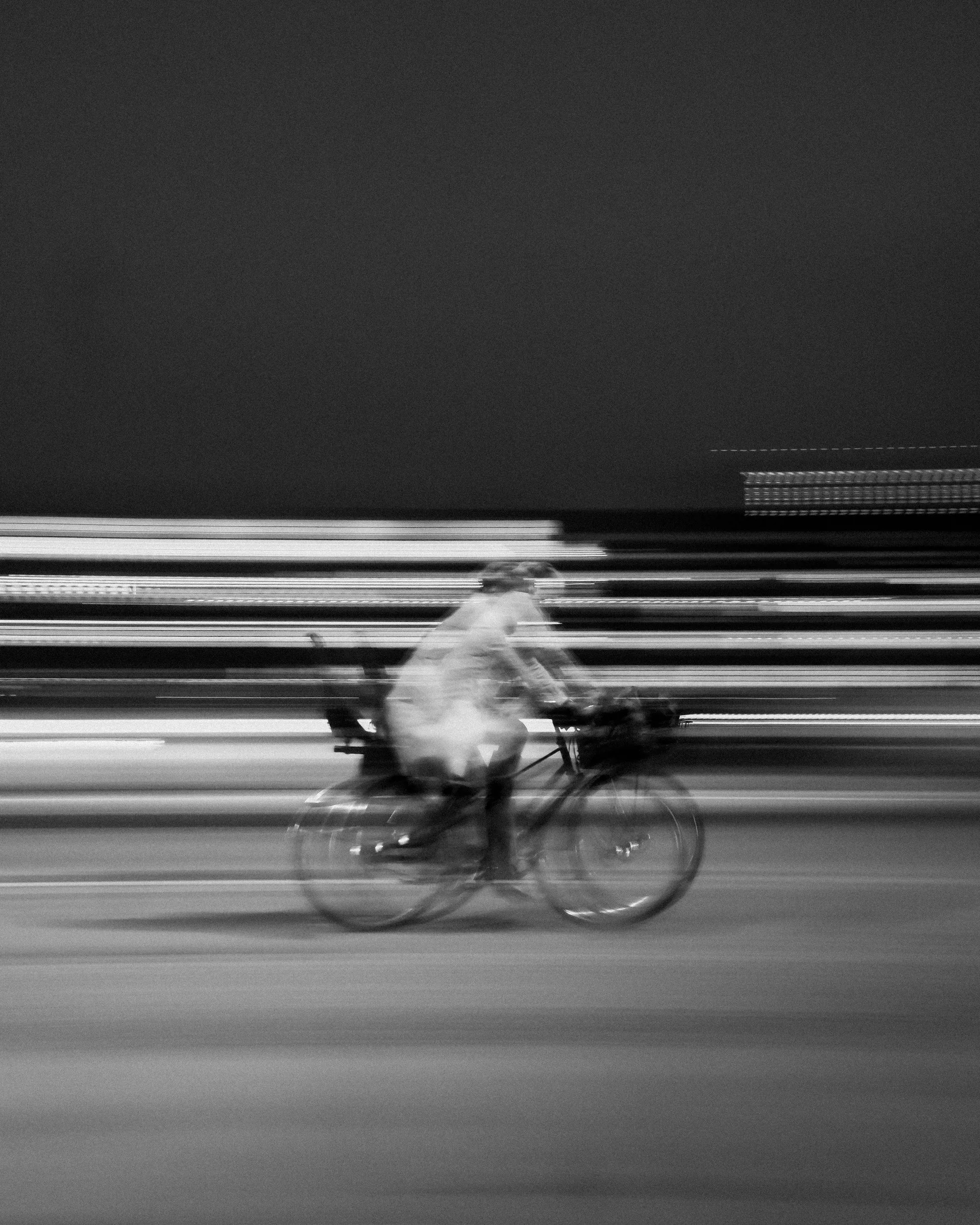 Black and white photo of a person riding a bicycle on a road, captured in motion blur.