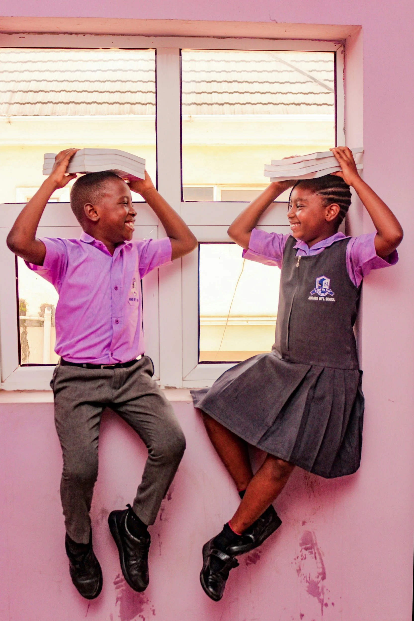 Two school children smiling and playing with books on their heads as they sit on a windowsill.