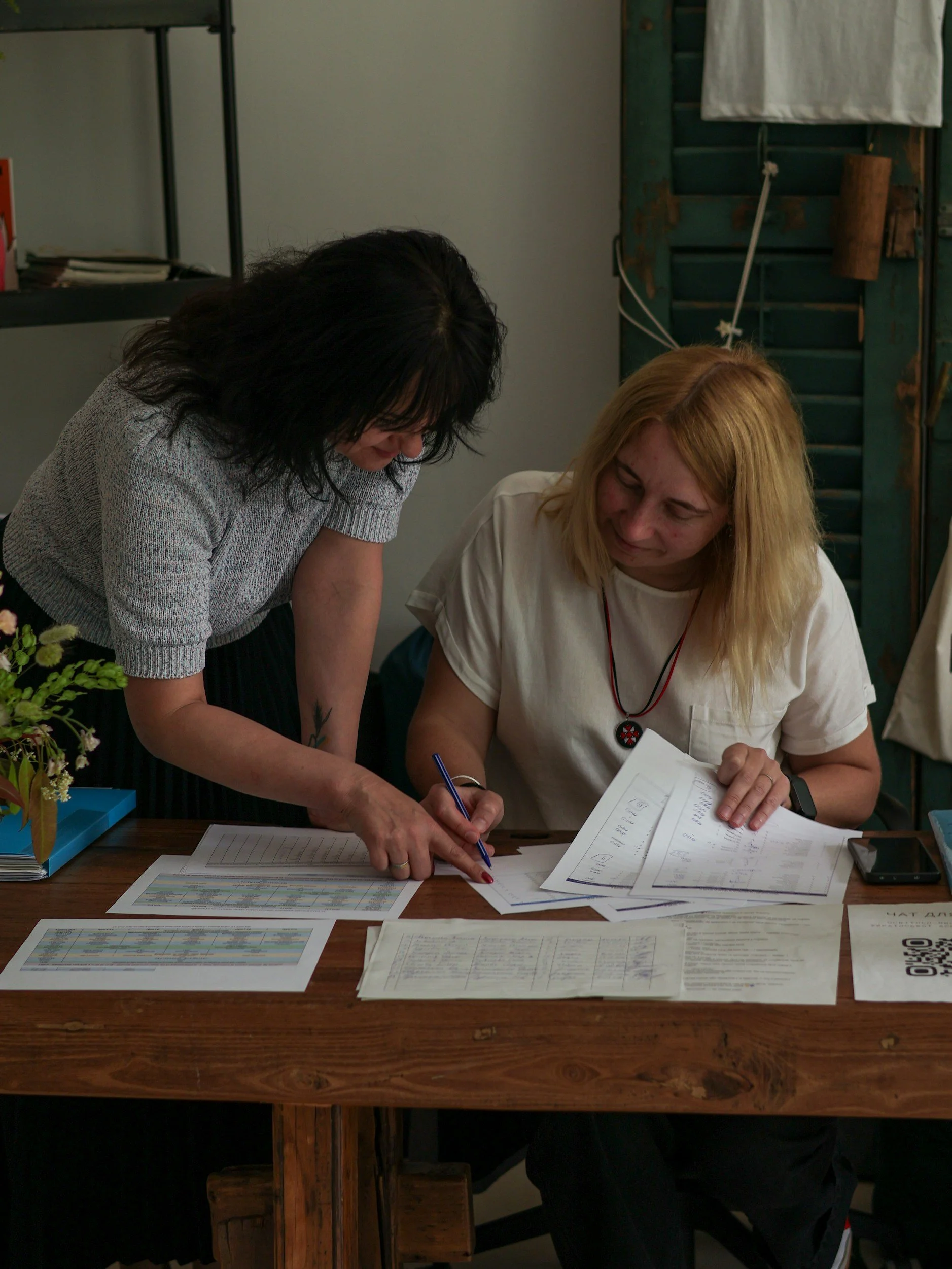 Two women are reviewing documents and charts at a wooden table, with one woman standing and pointing at the papers while the other sits and looks on.