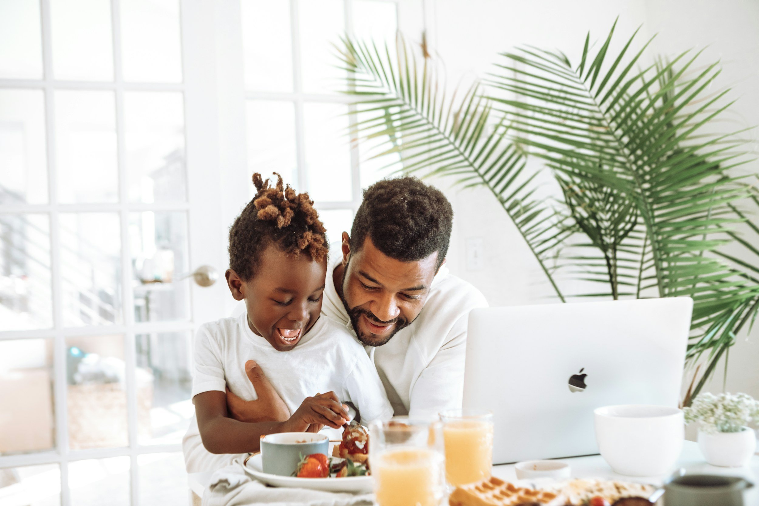 A father and his young child sharing a joyful moment at a breakfast table, surrounded by cakes and drinks, in a bright room with large windows and a green potted plant in the background.