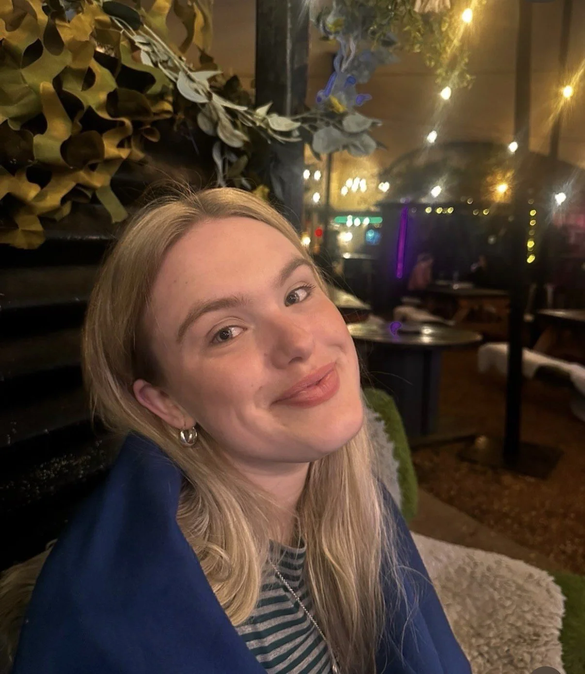 A young woman with blond hair smiling at the camera in a dimly lit restaurant or bar at night, with string lights and people in the background.