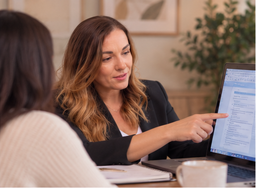 A woman with wavy brown hair points to a computer screen during a discussion at a meeting table in an office.