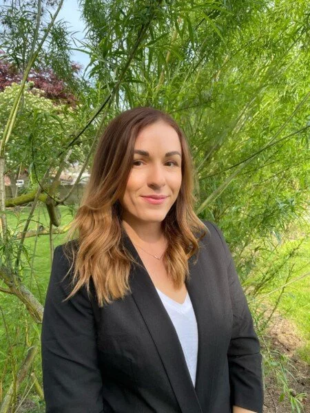 Woman with brown hair in a blazer outdoors surrounded by green trees and plants.