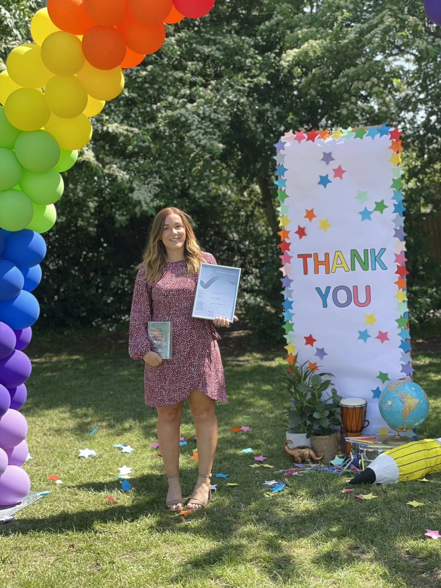 A young woman stands outdoors at a graduation celebration, holding a diploma and a glass award. She is smiling and dressed in a red and white dress. Colorful balloons arch over her, and a large white sign with multicolored stars and the words 'Thank You' is in the background. The scene is sunny with greenery and trees.