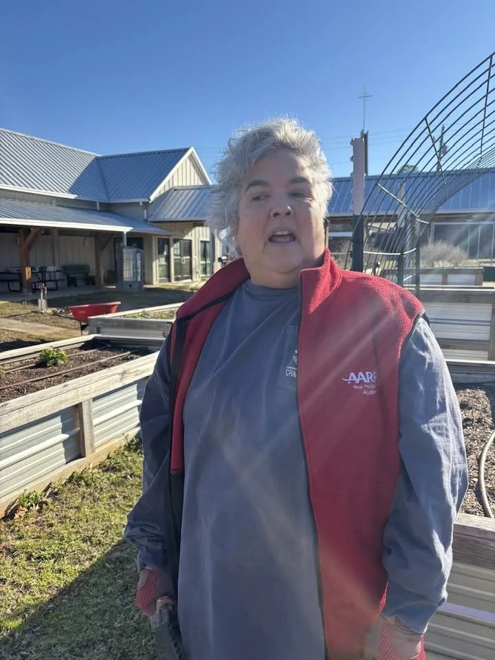 A person with short, gray hair wearing a red jacket over a blue shirt standing outdoors near a garden bed with a building in the background.