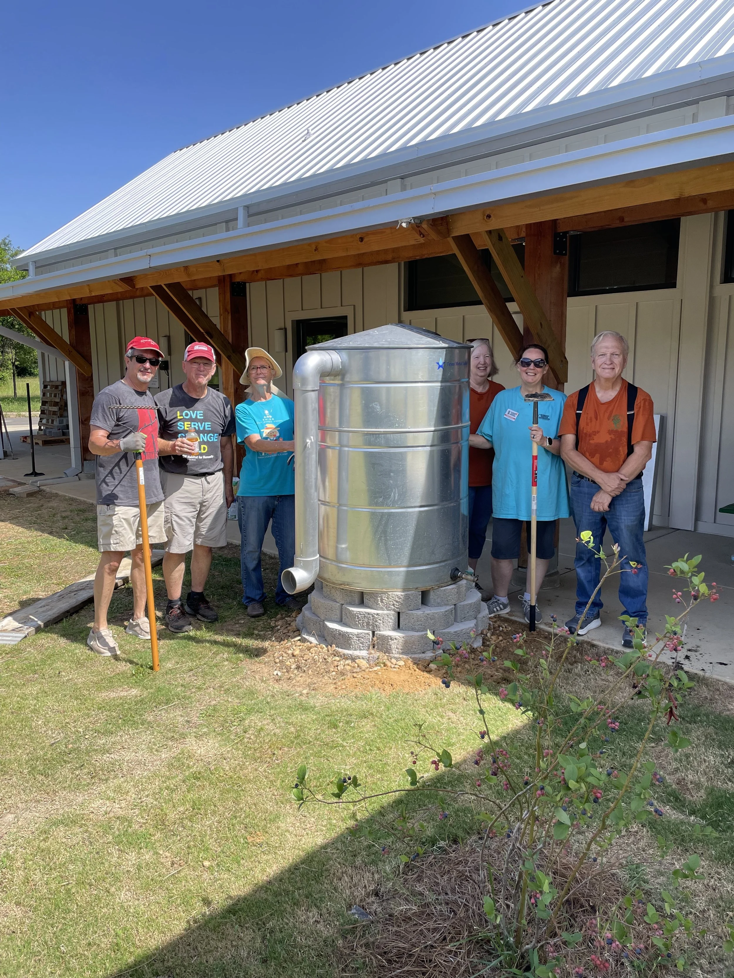Group of seven people standing outdoors around a metal water tank, some holding tools, in front of a building with a metal roof and wooden supports, under clear blue sky.