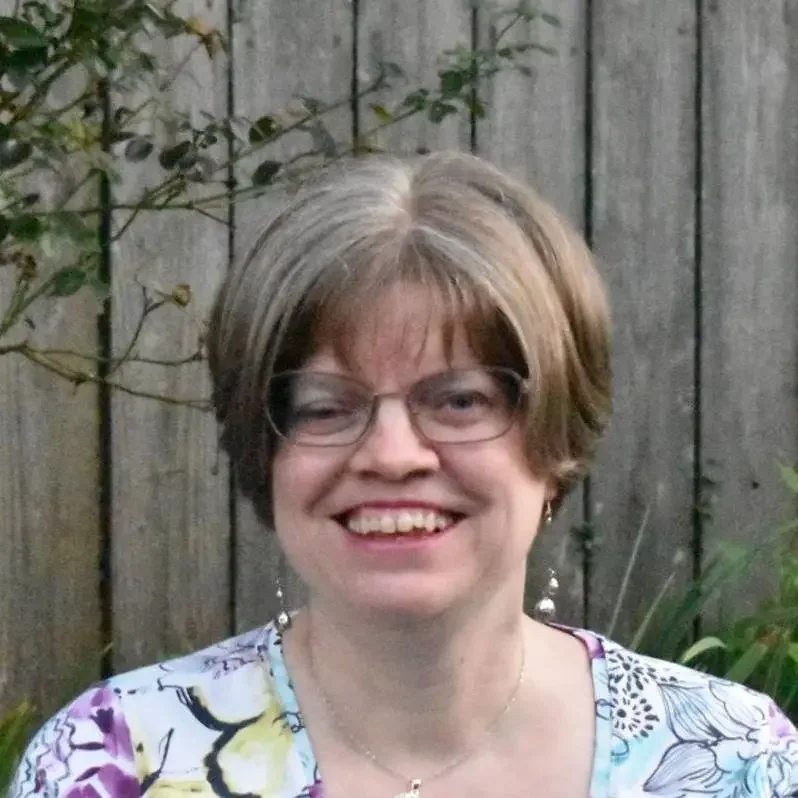 Smiling woman with short light brown hair and glasses, wearing a floral top and earrings, outdoors with a wooden fence and green plants in the background.