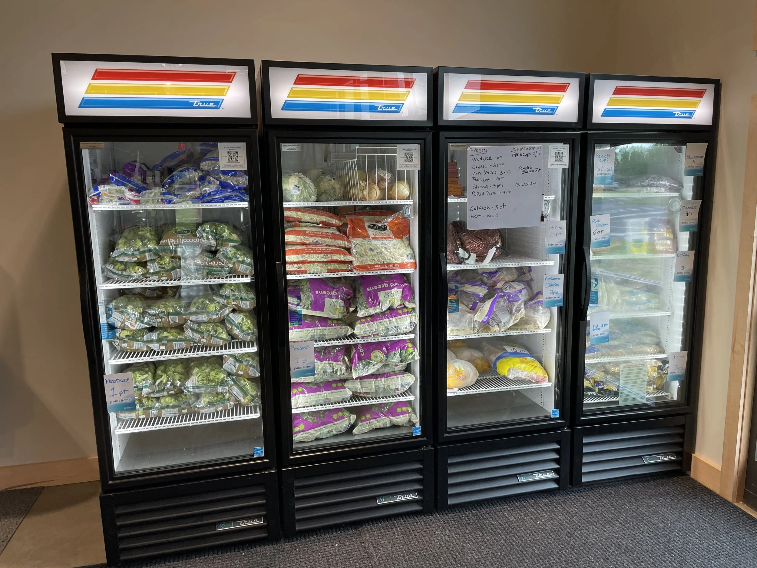 Four glass-front refrigerated display cases filled with frozen vegetables, meats, and other food items. Handwritten notes are attached to the middle and right cases, listing various food products and prices.