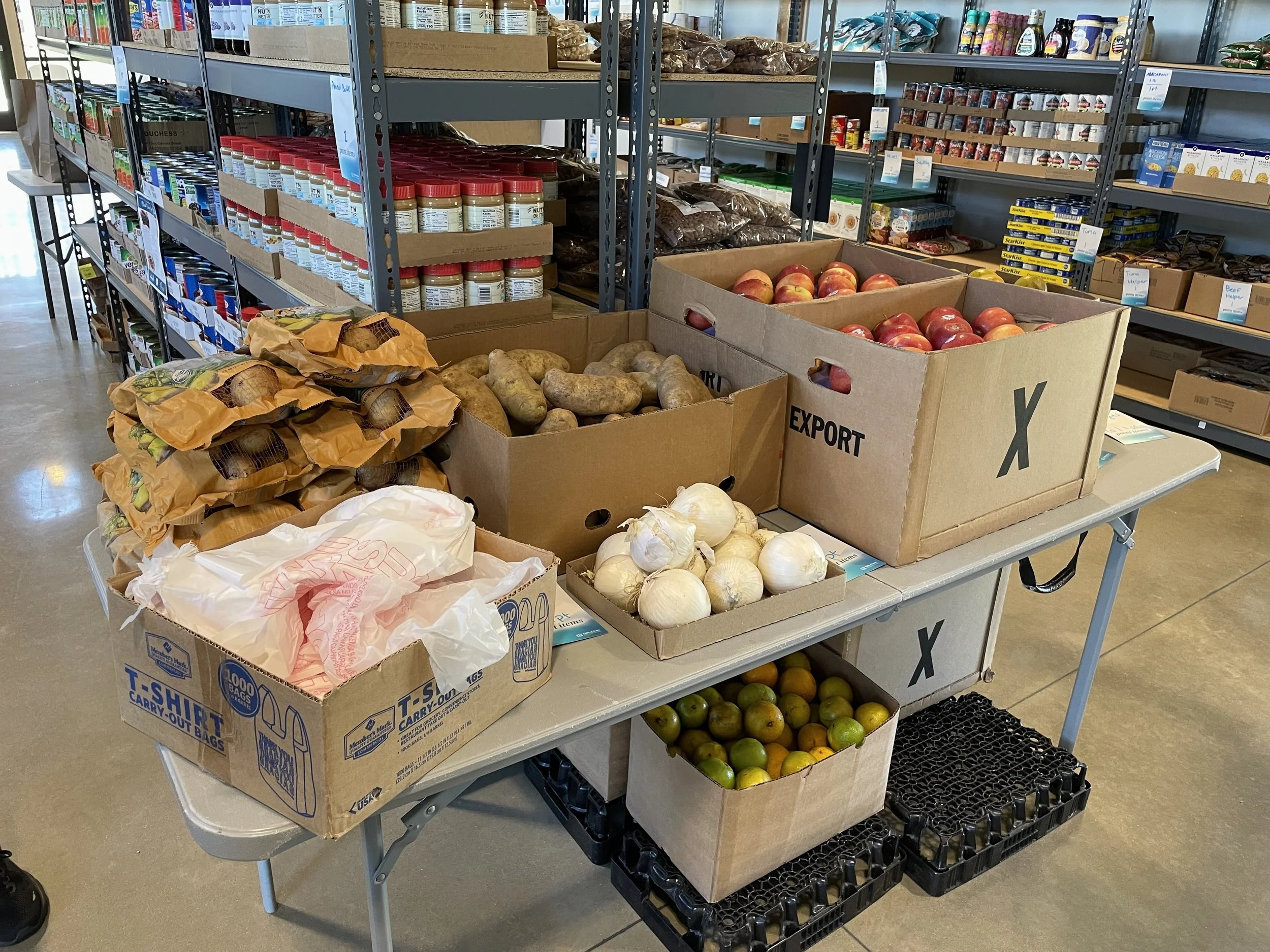 Table with boxes of produce including onions, garlic, potatoes, apples, and pears, with store shelves in the background.